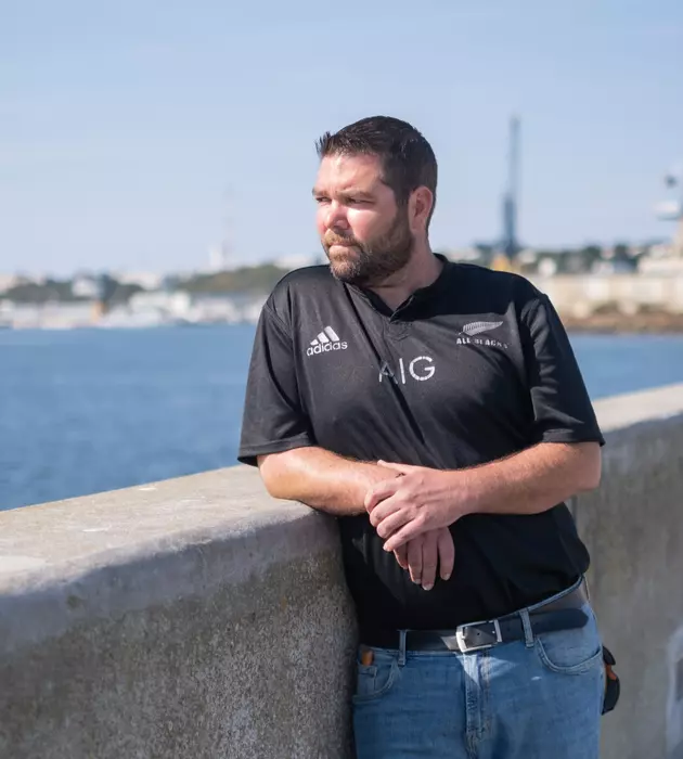 Homme accoudé à un mur de pierre avec la mer derrière lui qui regarde à sa droite, au loin.