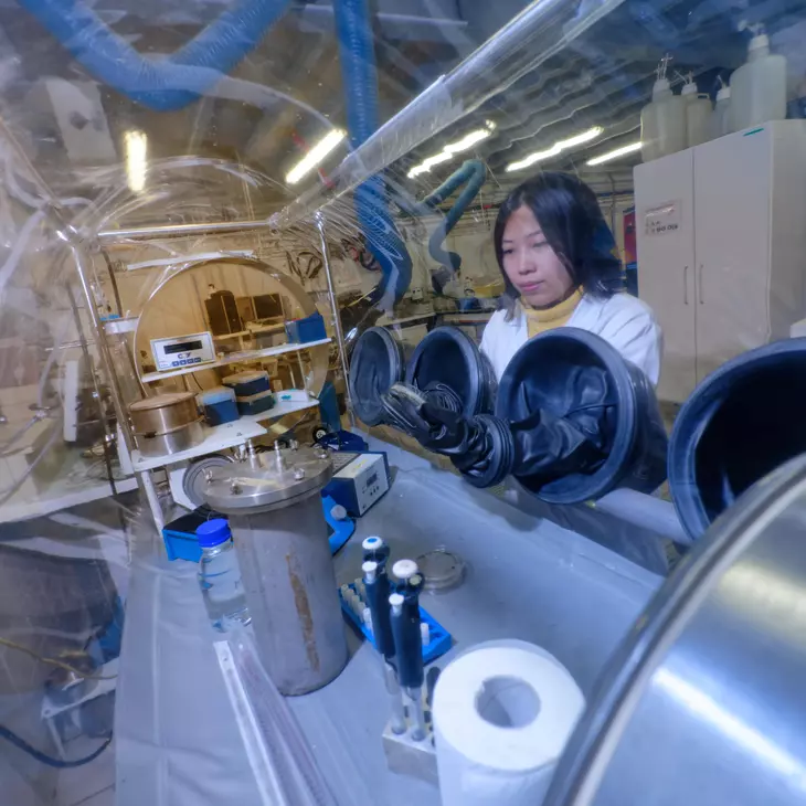 Personne en blouse blanche qui manipule des éléments dans un espace confiné (grand tube transparent).